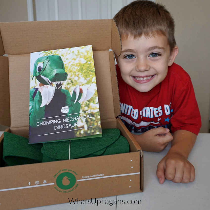 little boy smiling and posing with Kiwi Crate box featuring a chomping mechanical dinosaur kit that helps you make a 3d dinosaur head