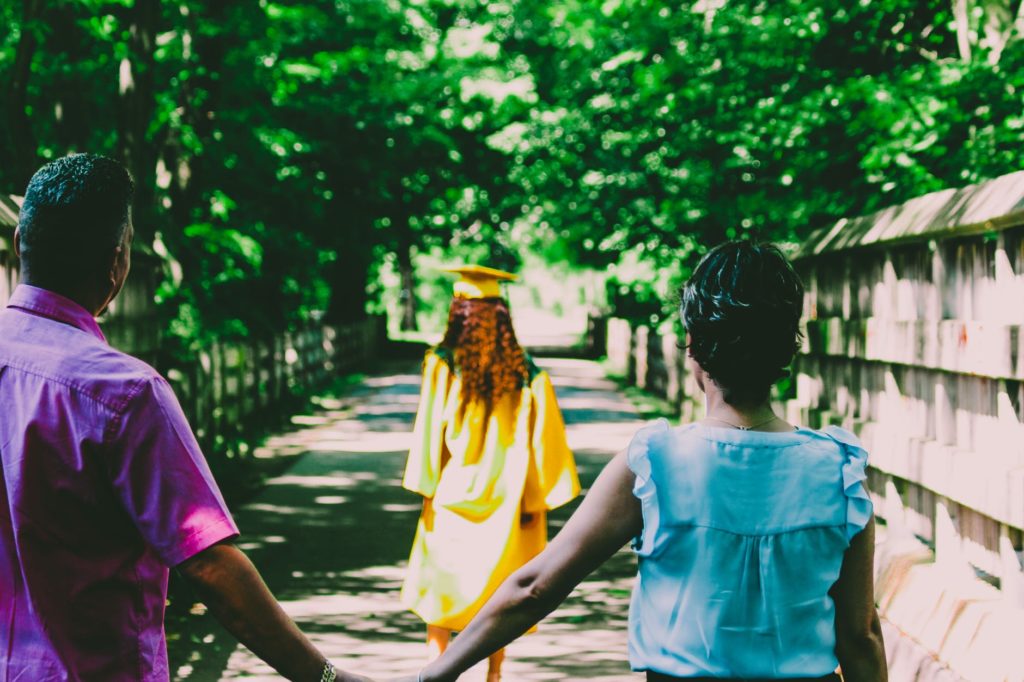 parental involvement in schools can help improve children's outcomes academically, shown by a husband and wife holding hands, looking after their daughter with a yellow graduation gown and cap on.